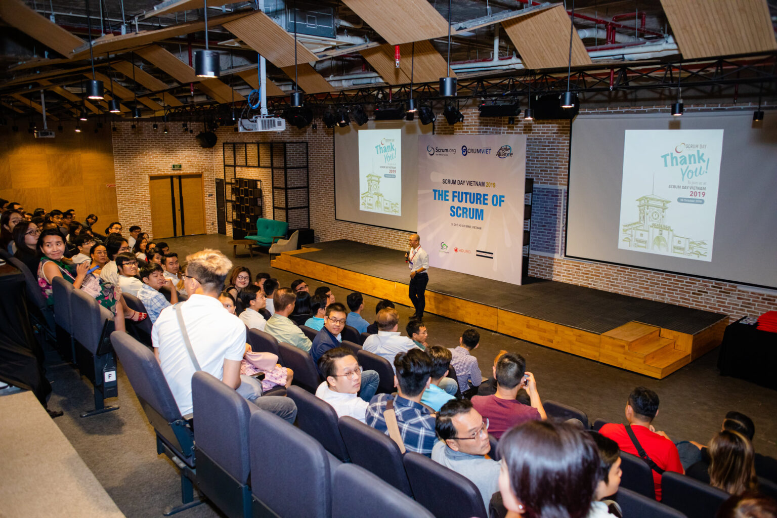 Khán phòng Scrum Day Vietnam 2019: diễn giả đứng trên sân khấu trước màn hình “The Future of Scrum”, khán giả ngồi kín các hàng ghế